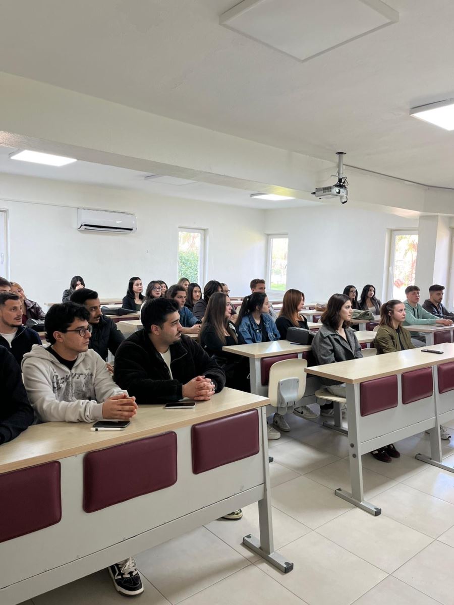 Bir üniversite sınıfında öğrenciler sıralarda oturarak yapılan sunumu dinlemektedir. Öğrenciler dikkatli bir şekilde ön tarafa bakarak toplantıyı takip etmektedir. Aydınlık bir sınıf ortamında gerçekleştirilen bu etkinlikte öğrencilerin çoğu not almak veya konuşmacıyı dinlemek üzere hazır durumdadır.